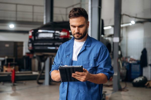 Modern digital tablet is in hands. Male mechanic is working in the car workshop.